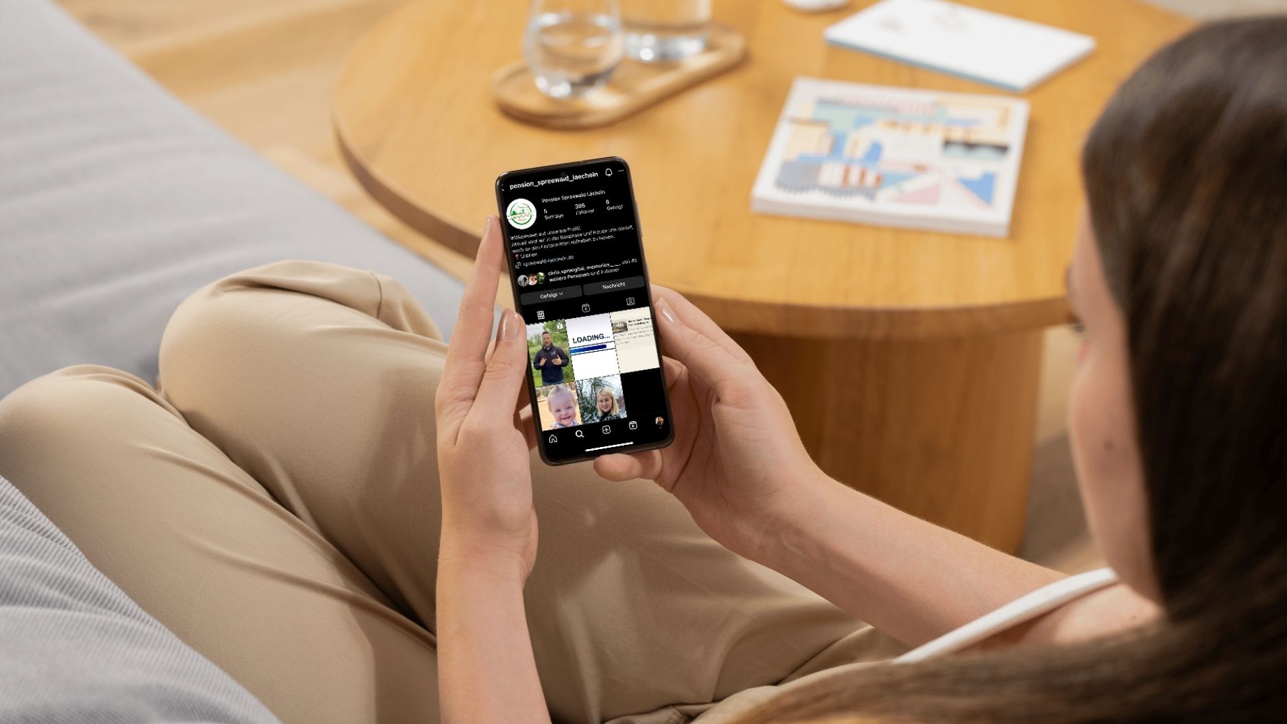 Eine Frau sitzt entspannt auf dem Sofa und hält ein Smartphone in der Hand, auf dem die Instagram-Seite der Pension Spreewald Lächeln geöffnet ist. Auf dem Bildschirm sind das Profilbild mit Logo, die Beschreibung sowie einige Beiträge der inklusiven Pension sichtbar.