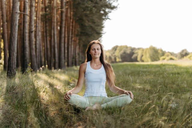 Eine Frau namens Eileen sitzt im Schneidersitz auf einer Wiese und praktiziert Yoga. Sie trägt ein helles, ärmelloses Oberteil und eine helle Yogahose, hat die Augen geschlossen und die Hände entspannt auf den Knien. Links steht ein dichter Kiefernwald, im Hintergrund öffnet sich eine sonnige, grüne Landschaft.